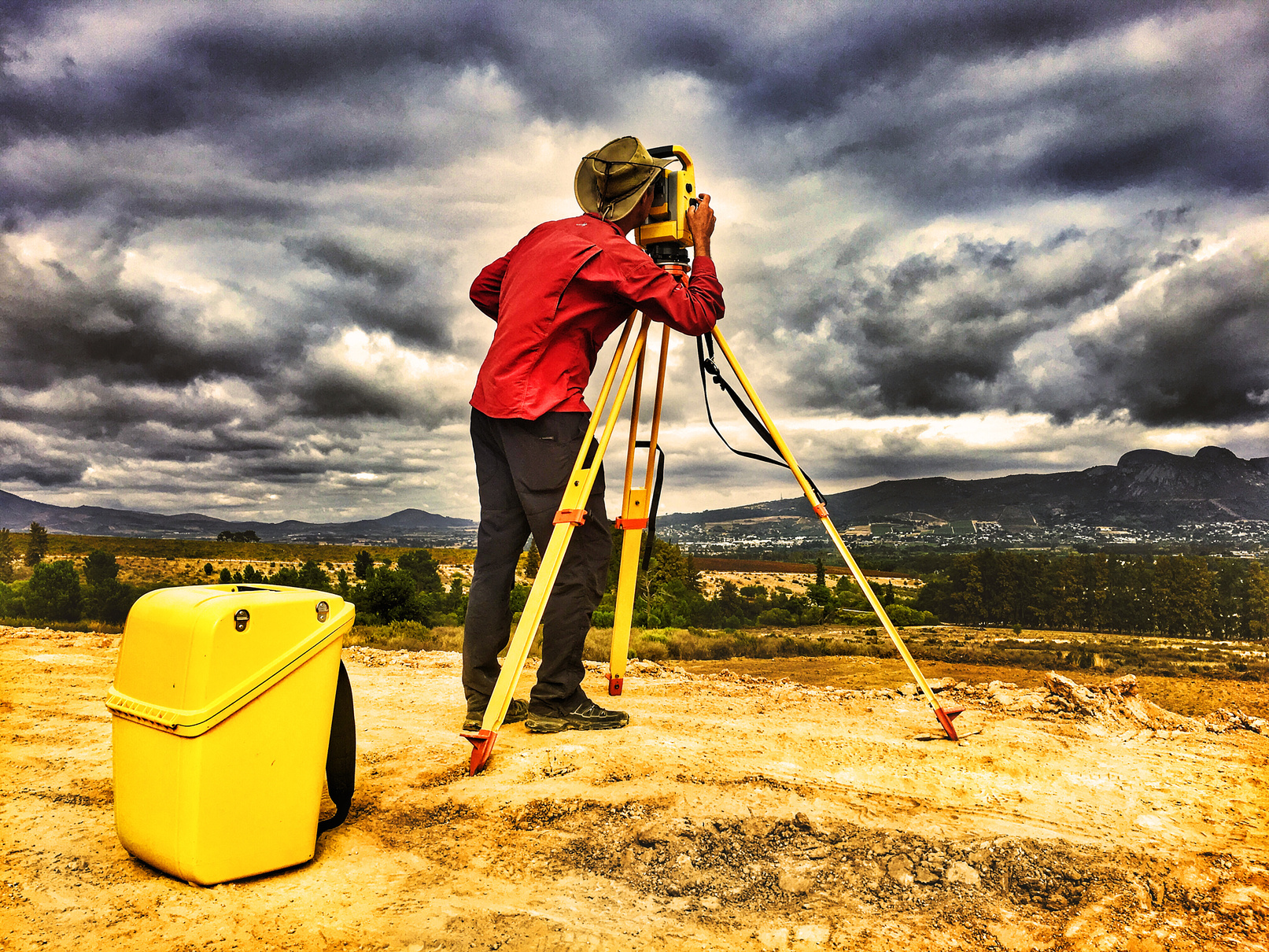 Land surveyor using total station for construction layout surveys during a earthworks project, dramatic stormy sky overhead, mountains in background